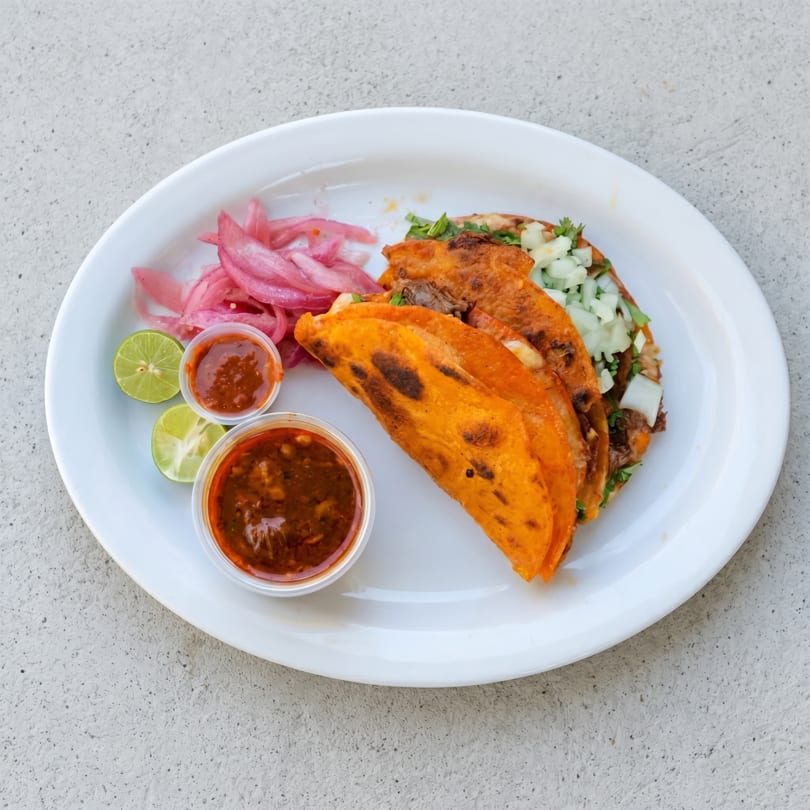 Three birria tacos with red onions, lime wedges, and two small cups of salsa on a white plate.